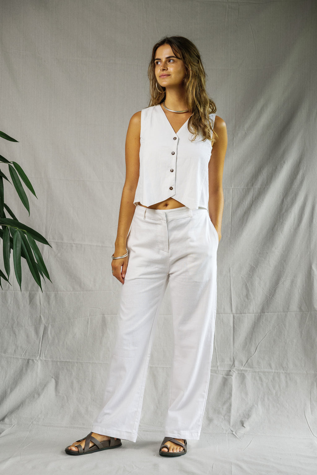 Woman wearing white breathable linen Odyssey Pants with high waist and relaxed fit paired with sleeveless buttoned white top .