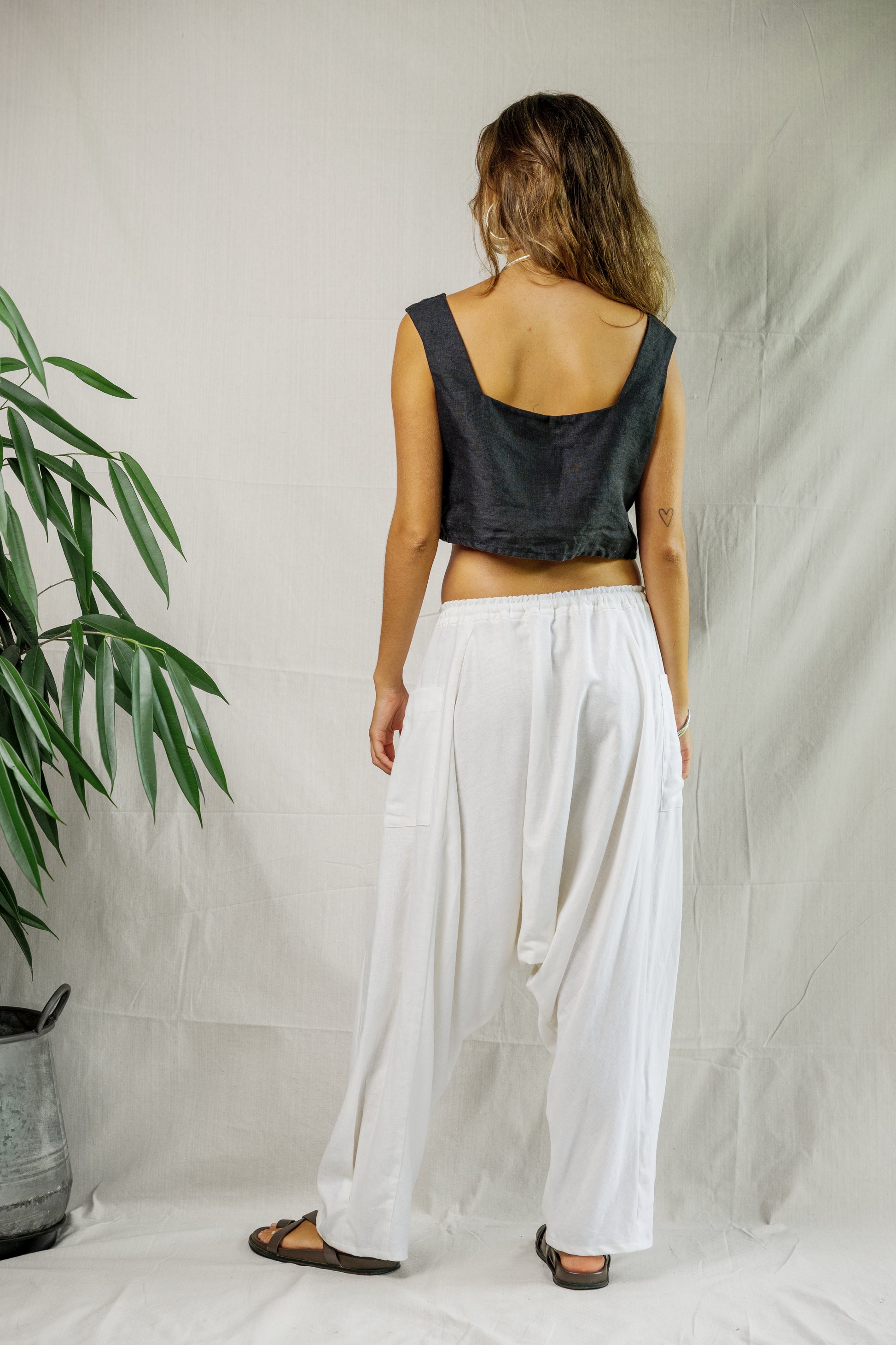 Woman wearing black linen sleeveless crop top and loose white breathable linen pants with sandals standing against white.