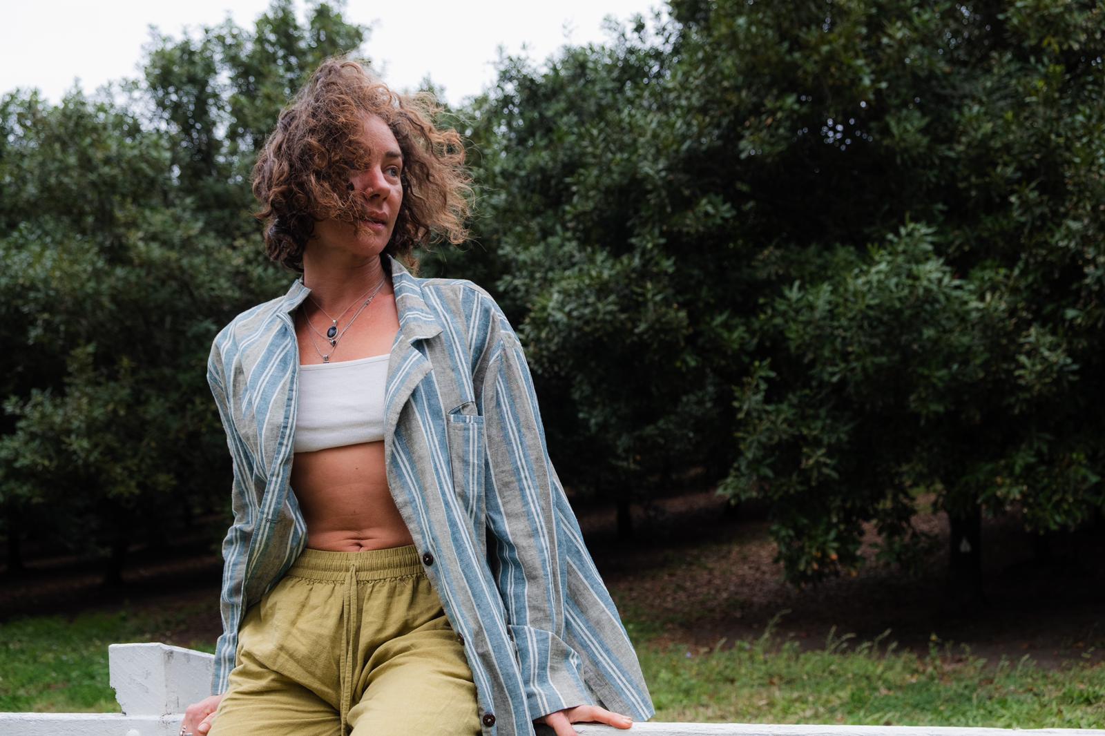 Person wearing a striped shirt over a crop top and high-waisted pants, standing outdoors with greenery in the background.