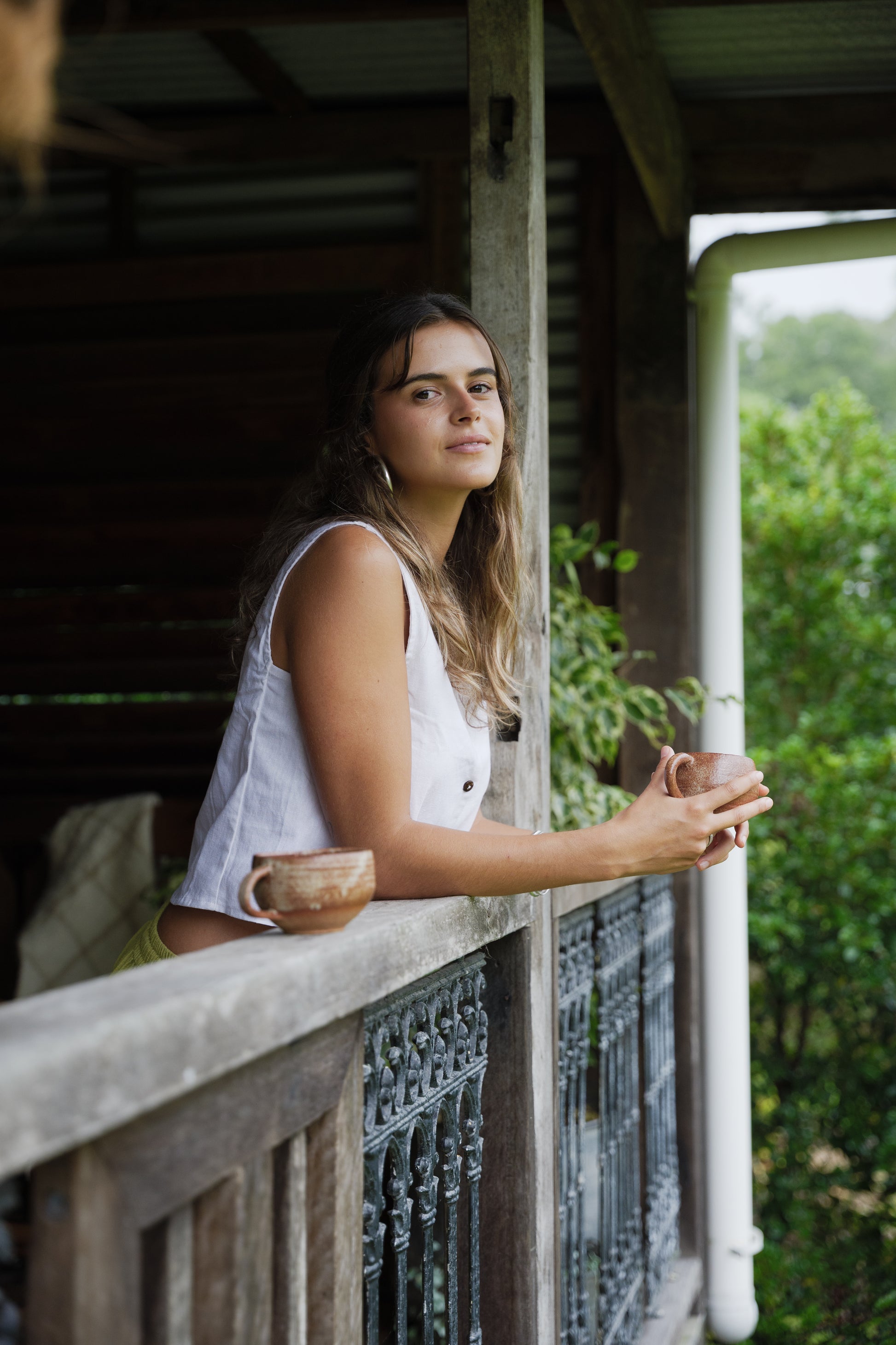 Sleeveless white linen vest with button front and relaxed fit worn by model leaning on balcony railing.