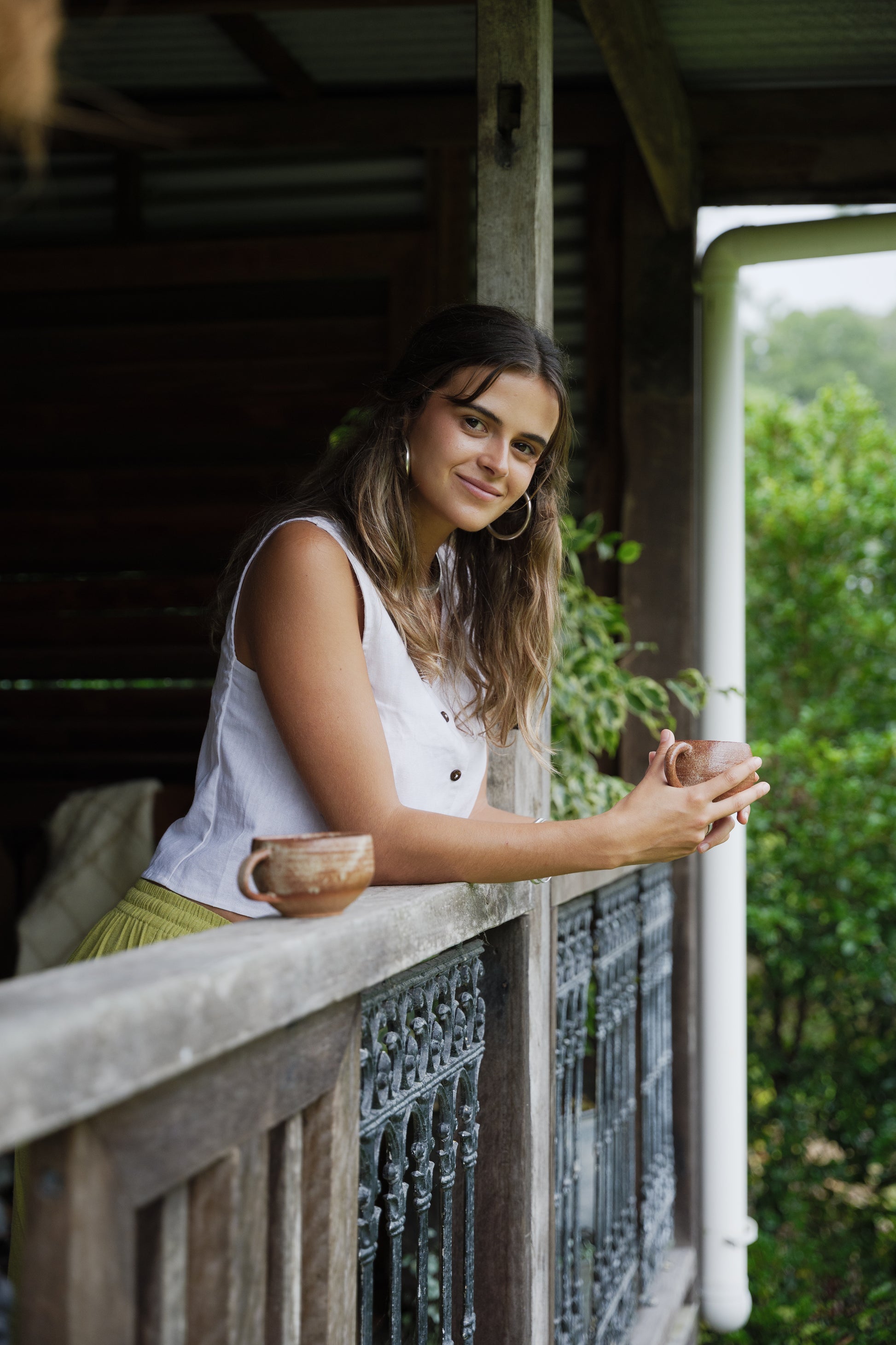White sleeveless linen vest with button front and lightweight breathable texture worn by model leaning on rustic wooden.