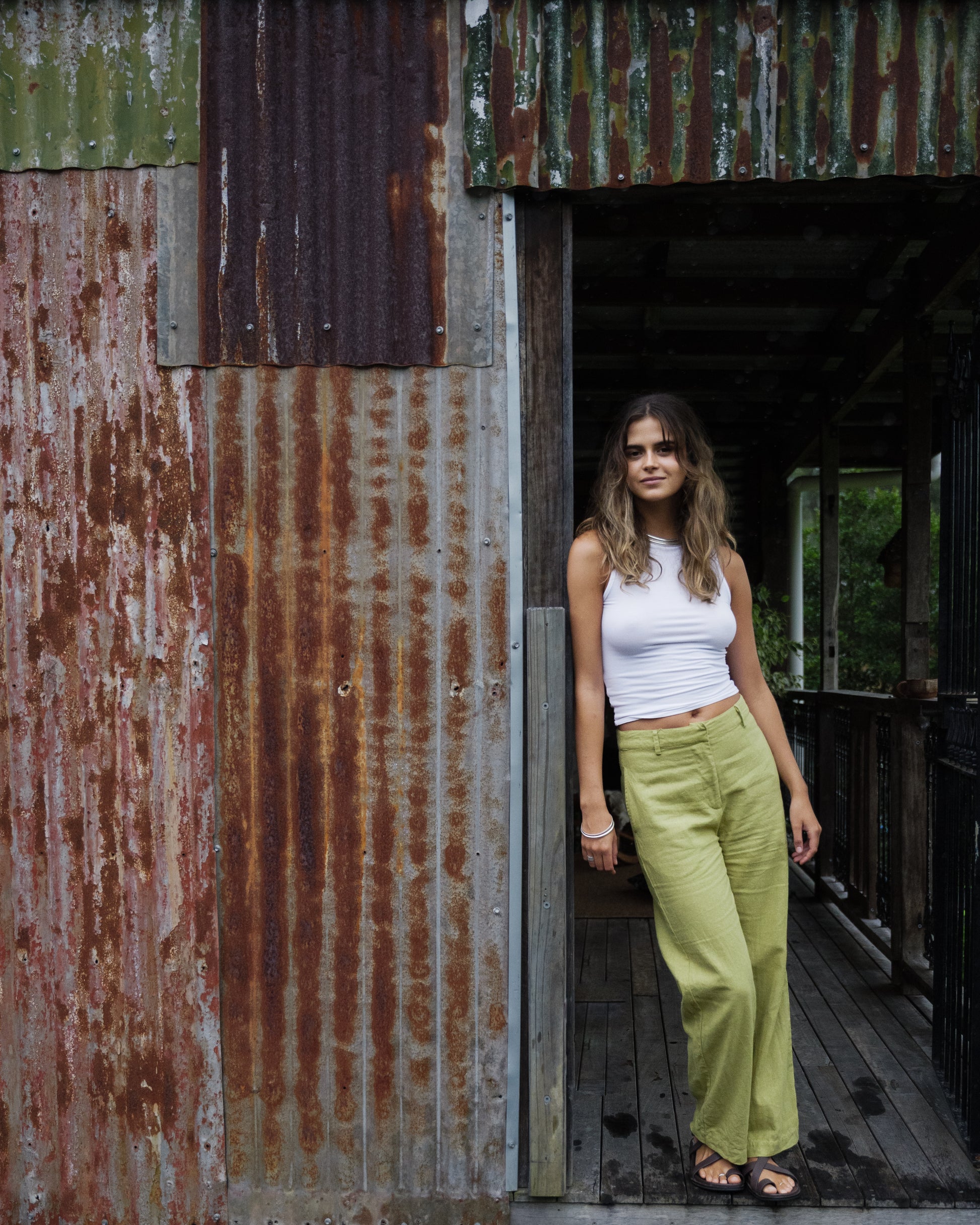 Woman wearing pistachio linen breezy straight-leg pants and white sleeveless top standing against rustic metal wall.