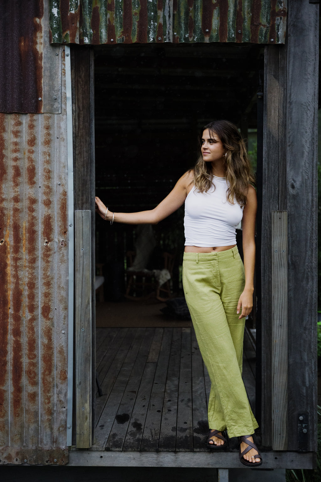 Woman wearing pistachio linen wide-leg Odyssey Pants and white sleeveless top standing in rustic doorway.