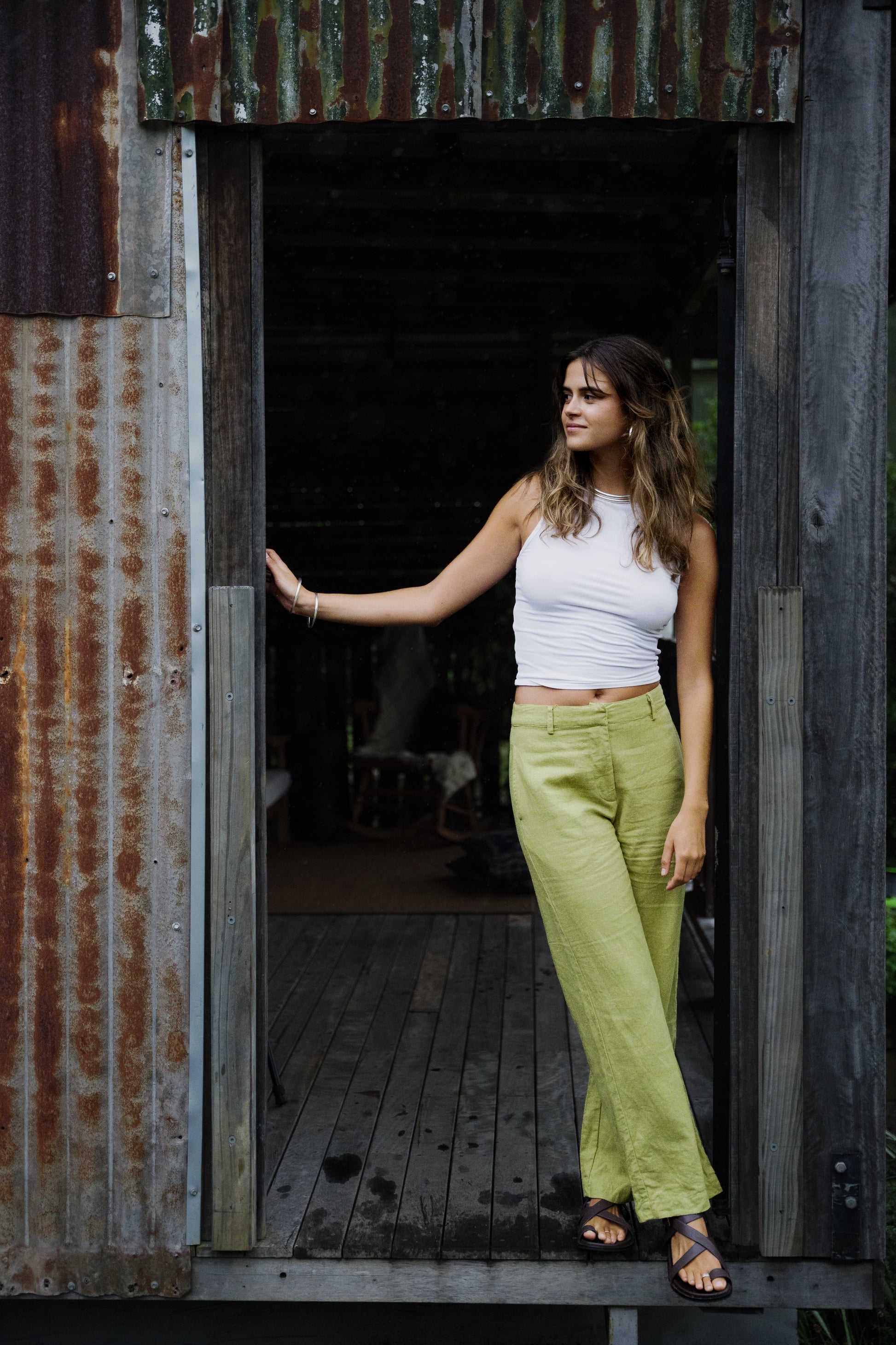 Woman wearing pistachio linen wide-leg Odyssey Pants and white sleeveless top standing in rustic doorway.