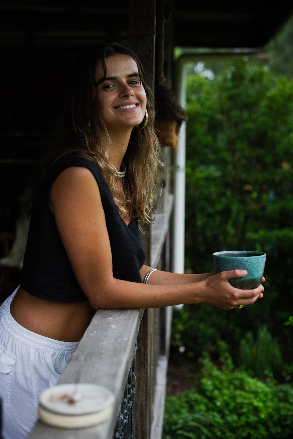 Woman wearing a black sleeveless crop top and white linen pants, holding a teal cup while leaning on a wooden railing.