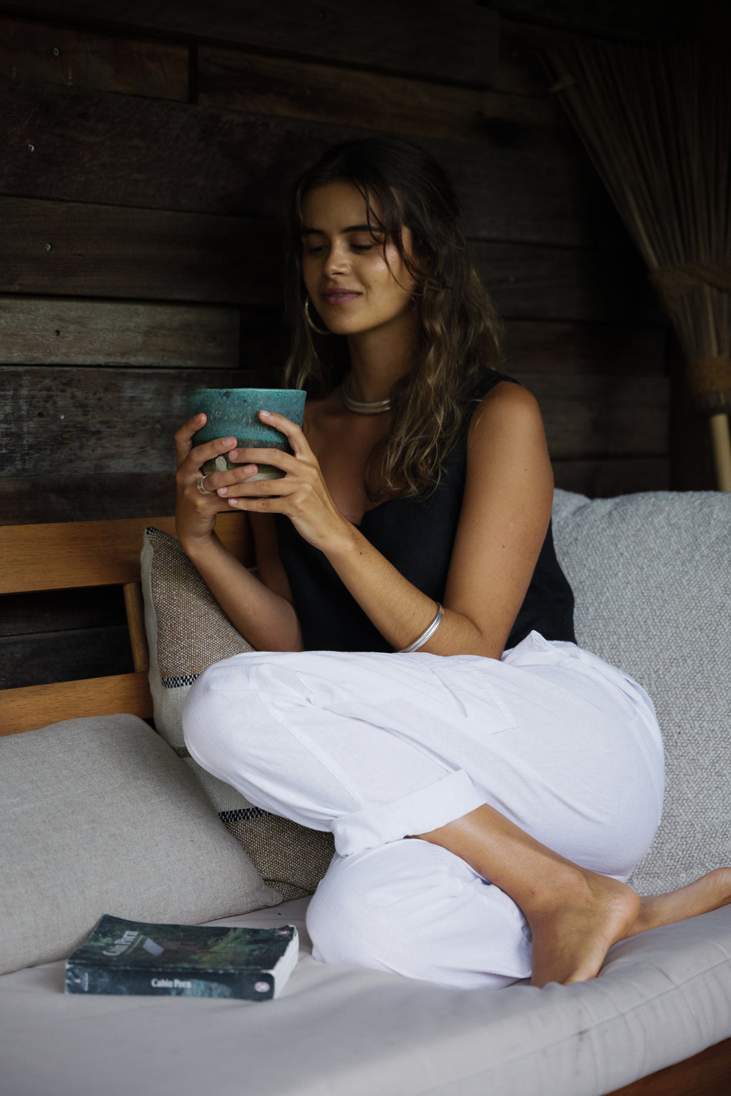 Woman seated cross-legged on a gray cushion wearing white linen cargo pants and a black tank top holding a teal mug.