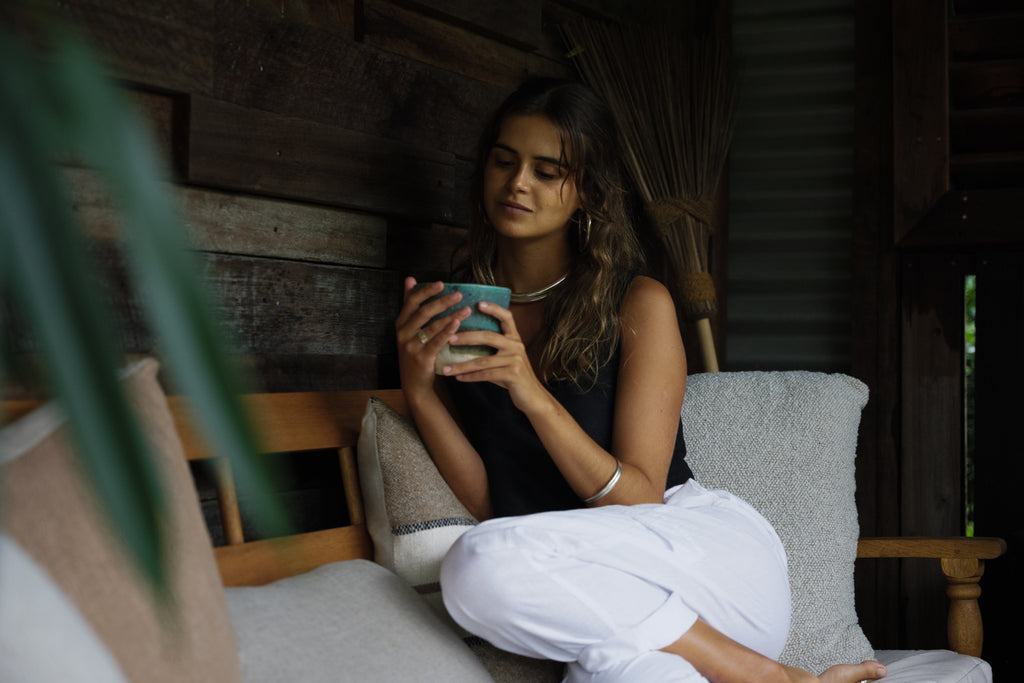 Woman wearing white linen Nomad Cargo Pants sitting cross-legged on a wooden bench holding a cup.