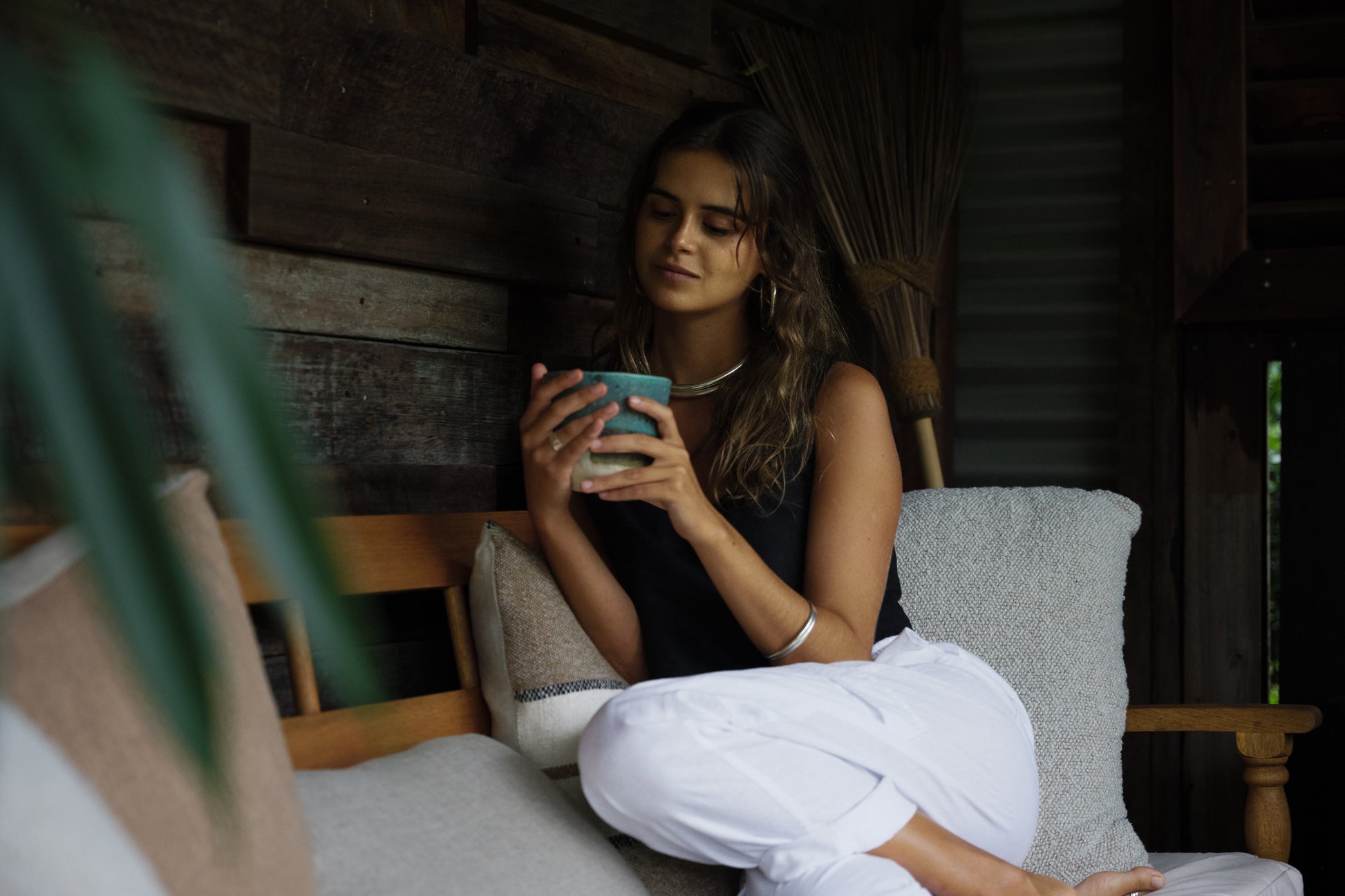 Woman wearing white linen Nomad Cargo Pants sitting cross-legged on a wooden bench holding a cup.