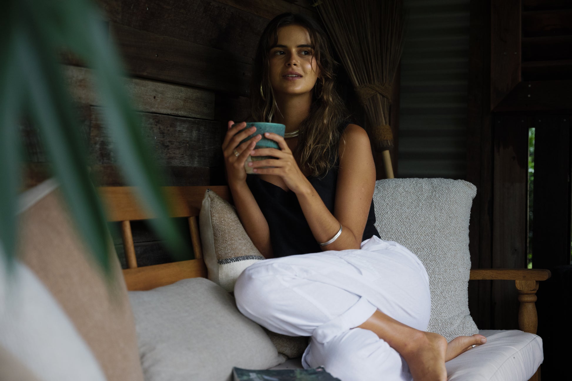 Woman wearing black sleeveless top and white linen cargo pants sitting on cushioned wooden bench holding a blue mug.