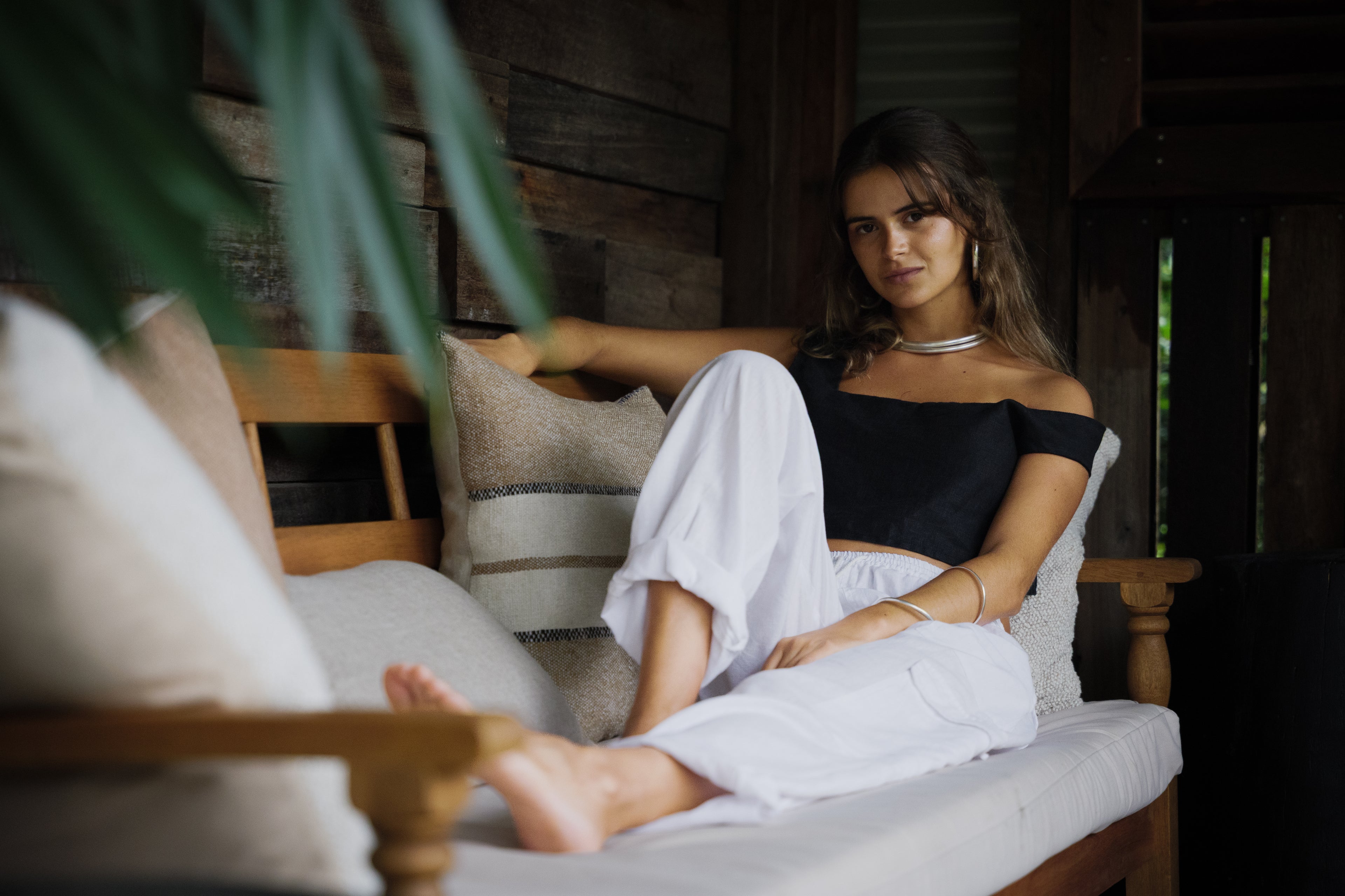 Woman sitting on wooden bench wearing black off-shoulder crop top and white loose linen pants.