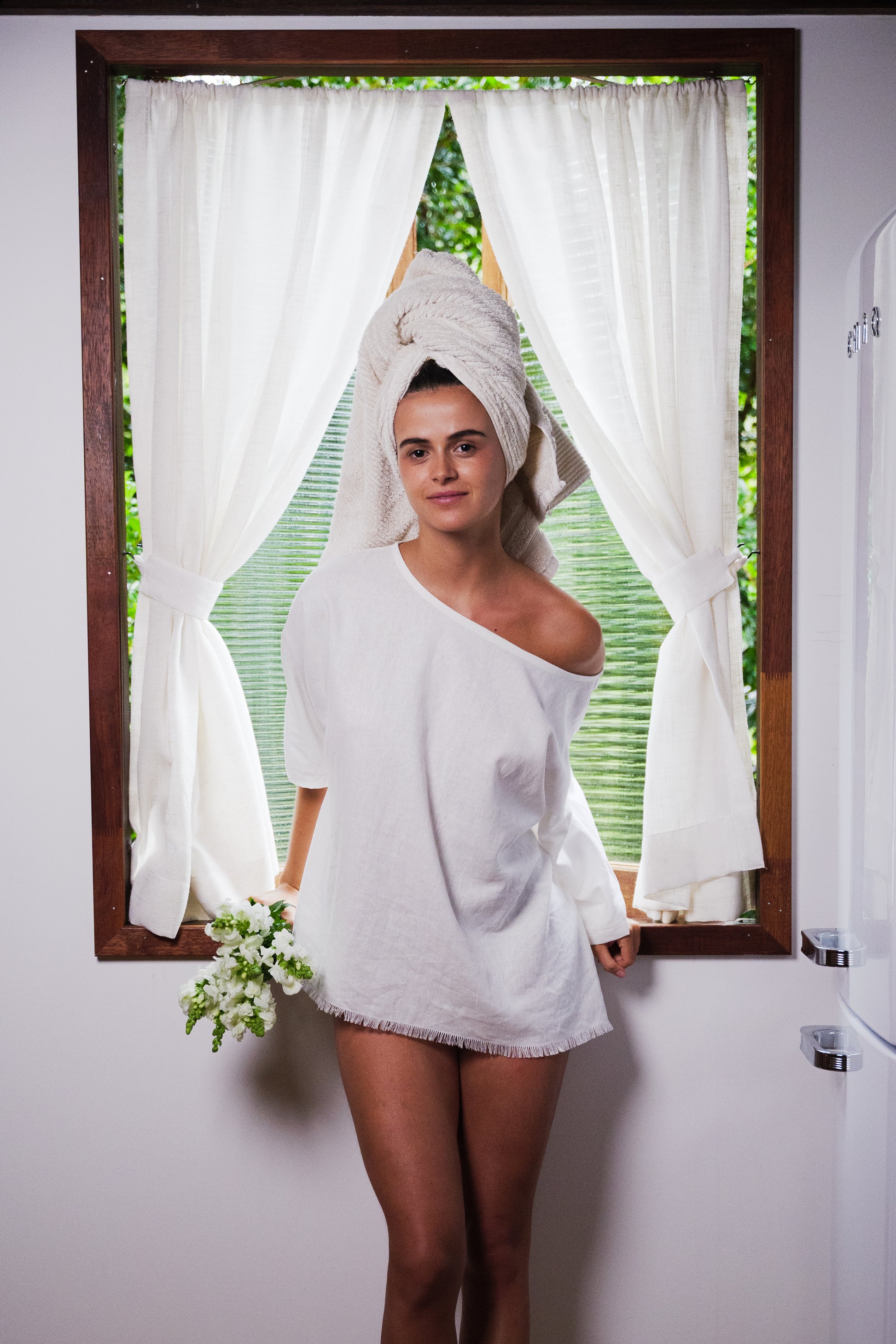 White linen off-shoulder beach tee worn with a towel wrapped on the head, holding small white flowers.