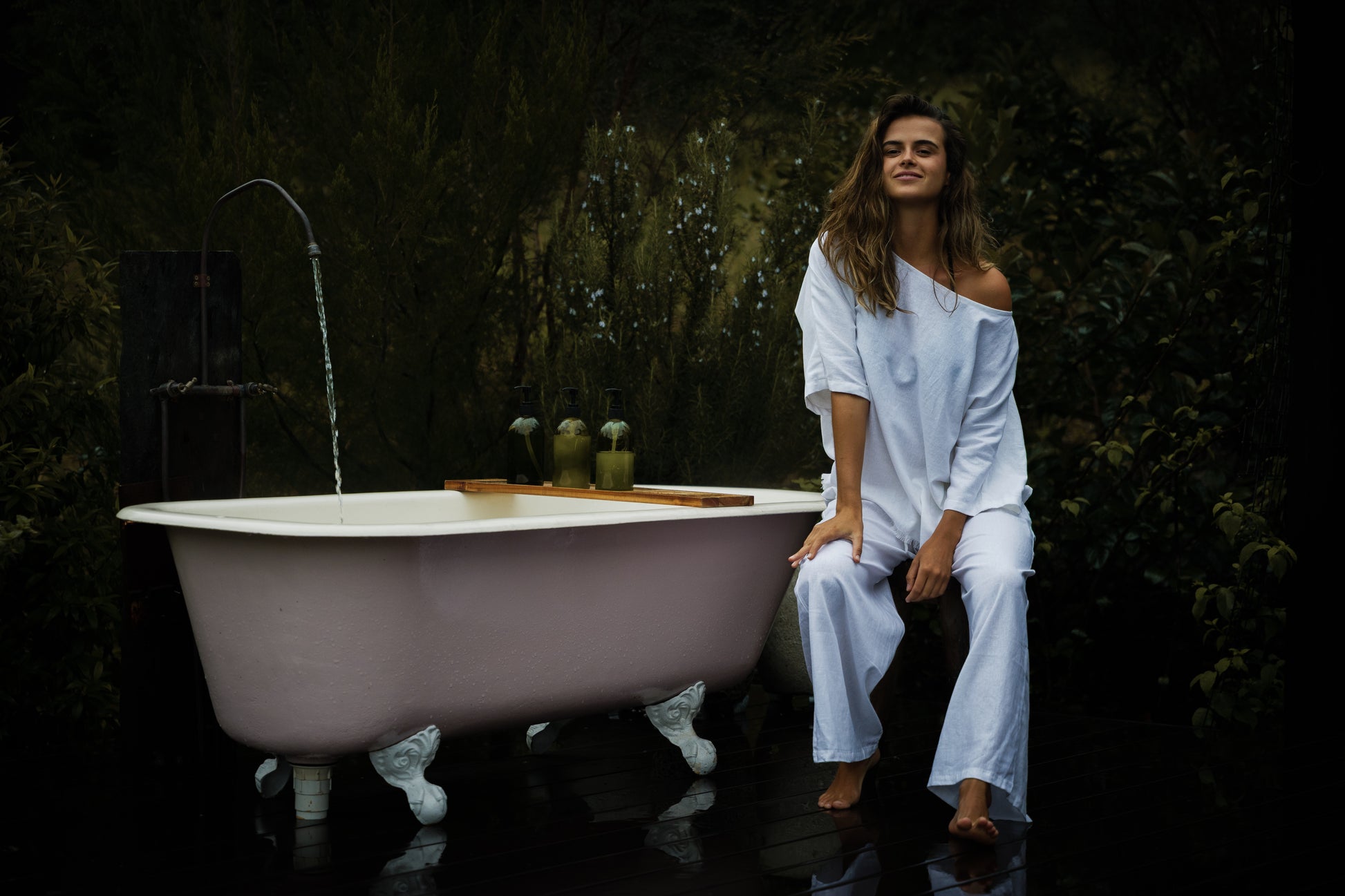 Woman wearing loose white linen Sol Pants and matching top standing barefoot next to clawfoot bathtub outdoors.
