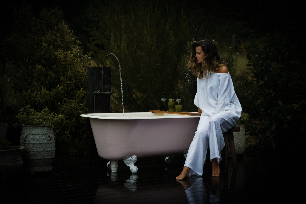 White breathable linen pants and matching long-sleeve top worn by woman sitting next to vintage clawfoot bathtub outdoors.