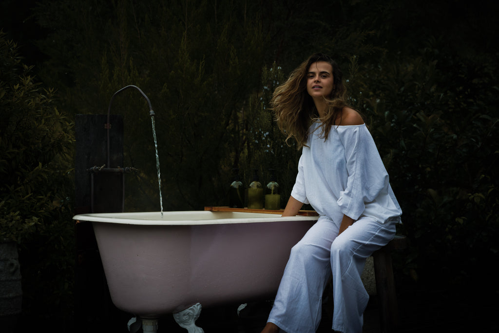 Loose white linen pants paired with a matching off-shoulder top, worn by a seated woman next to a vintage clawfoot bathtub.