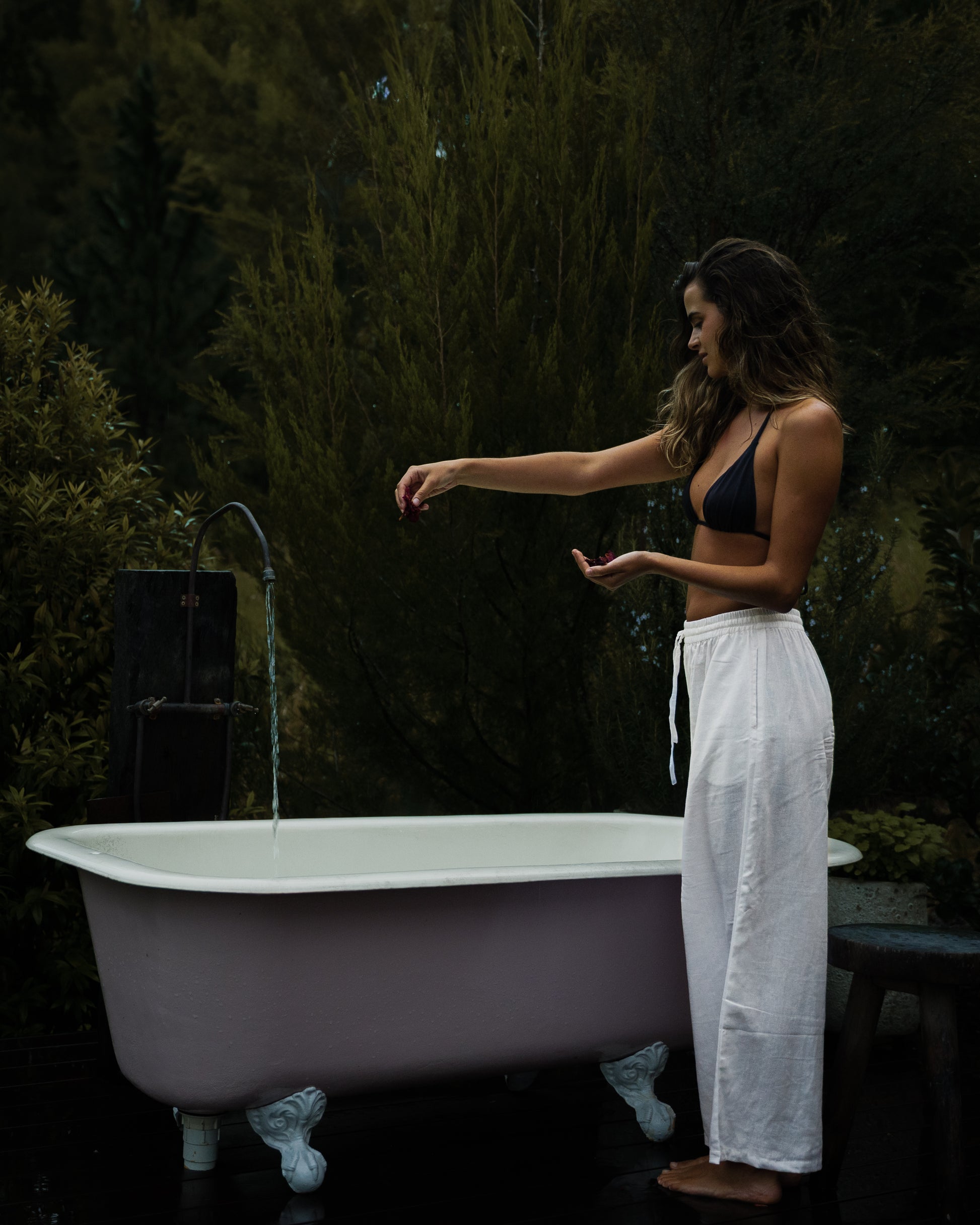 Woman wearing white linen pants and black crop top standing next to freestanding bathtub outdoors.