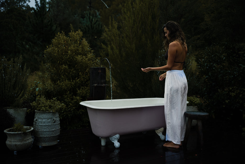 White linen Sol Pants with relaxed fit and cropped length worn by a woman standing beside a freestanding bathtub outdoors.