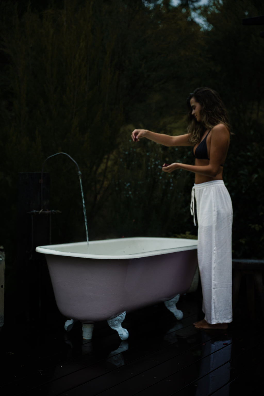Woman wearing white high-waisted linen pants with a tie waist, standing next to a pink clawfoot bathtub outdoors.