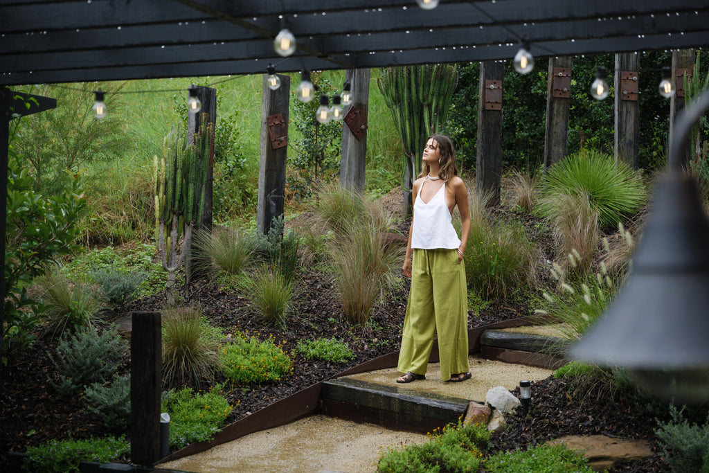 Woman wearing jungle green wide-leg linen pants and white sleeveless top in outdoor garden setting.