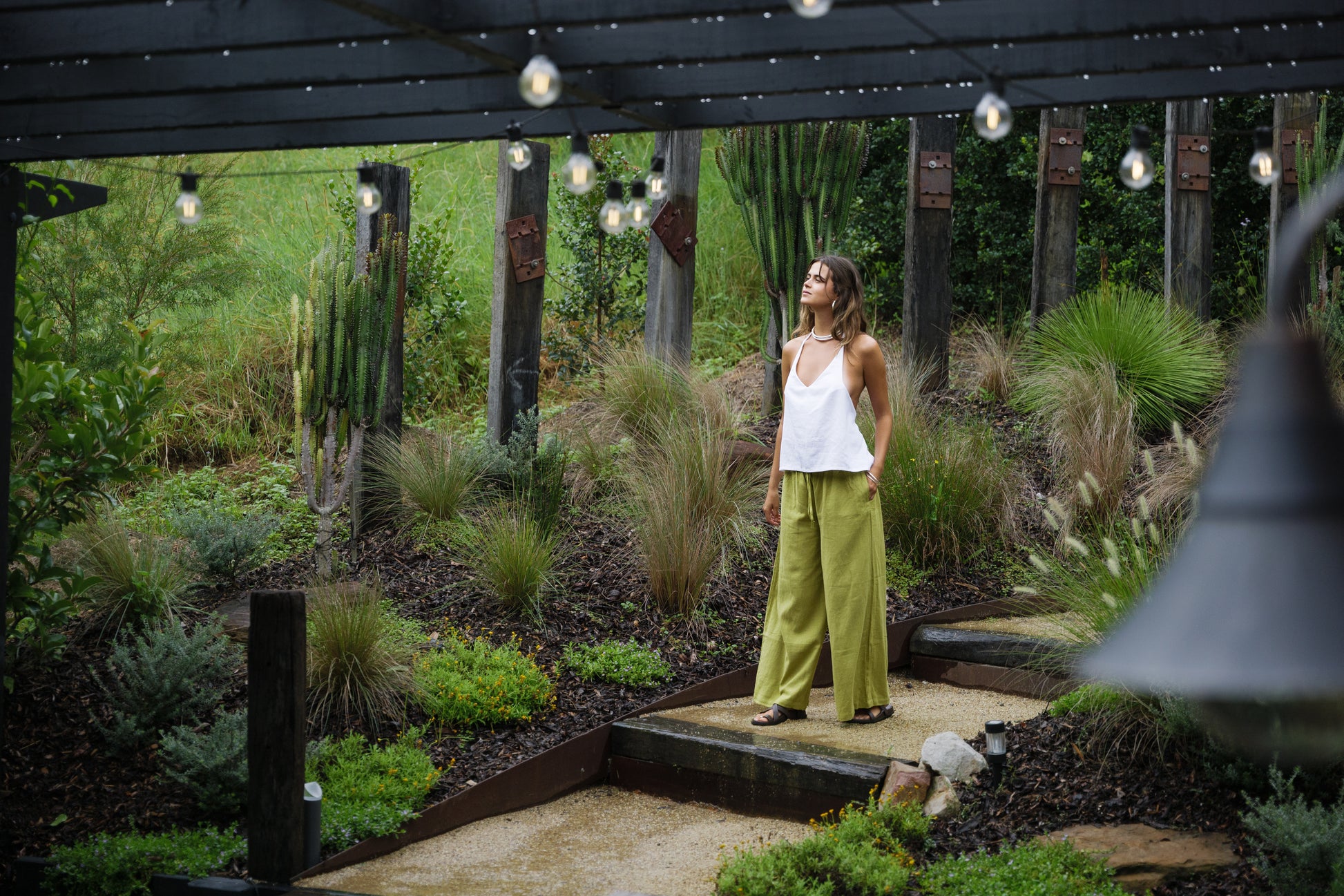 Woman wearing jungle green wide-leg linen pants and white sleeveless top in outdoor garden setting.
