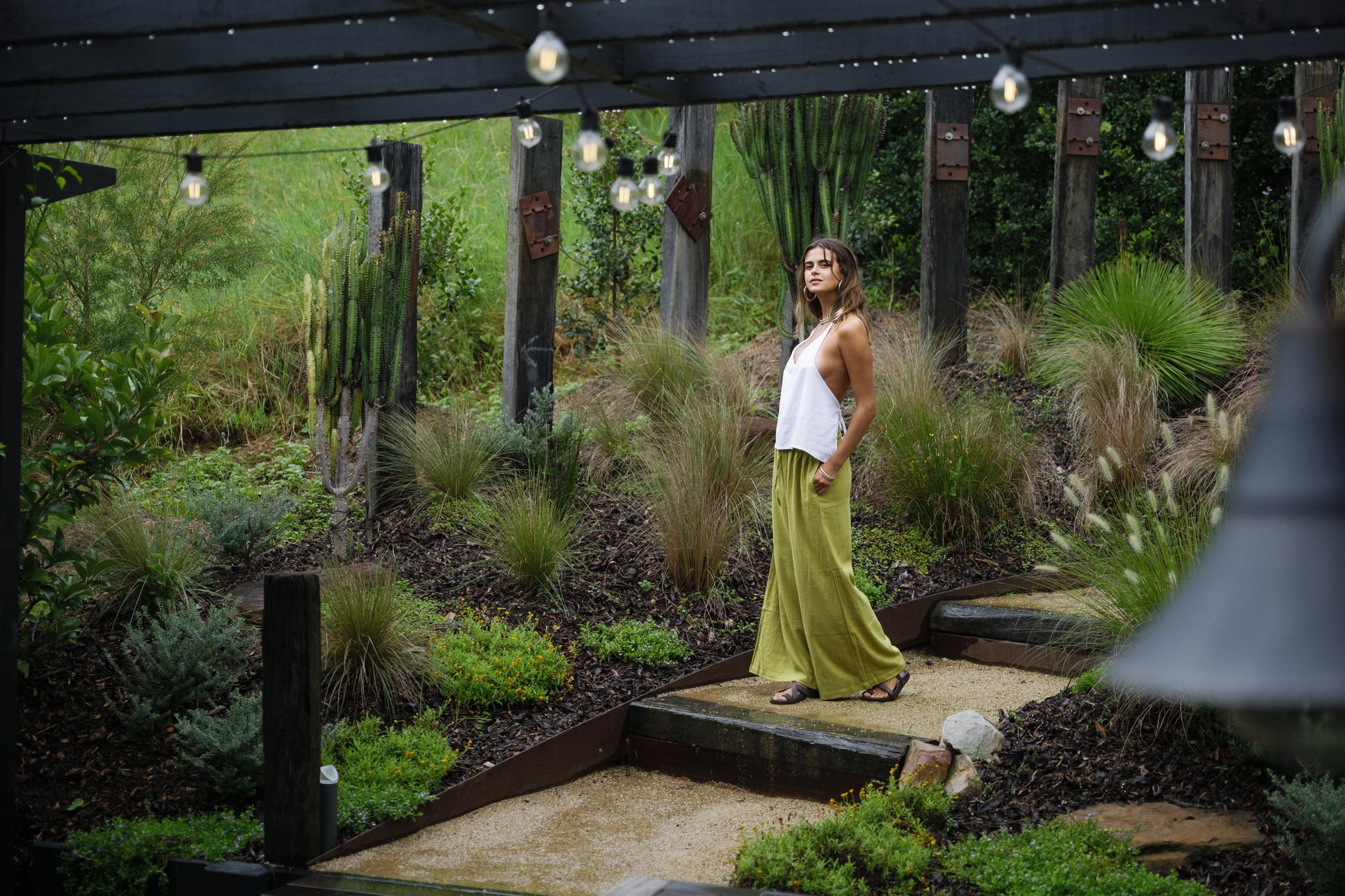 Woman wearing flowing jungle green linen pants and white sleeveless top in a lush garden setting.