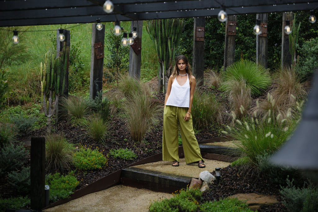Woman wearing breathable jungle green linen Sol Pants with white tank top in outdoor garden setting.