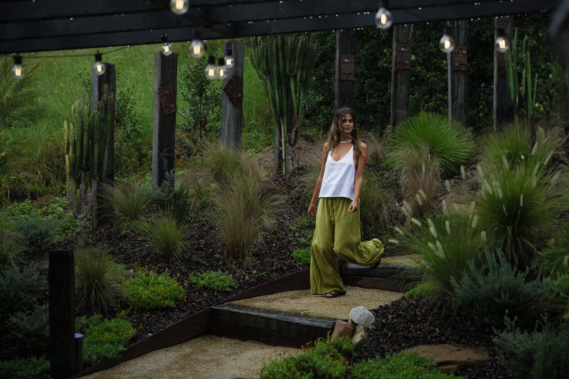 Woman wearing loose jungle green linen pants with a white sleeveless top walking outdoors at dusk.