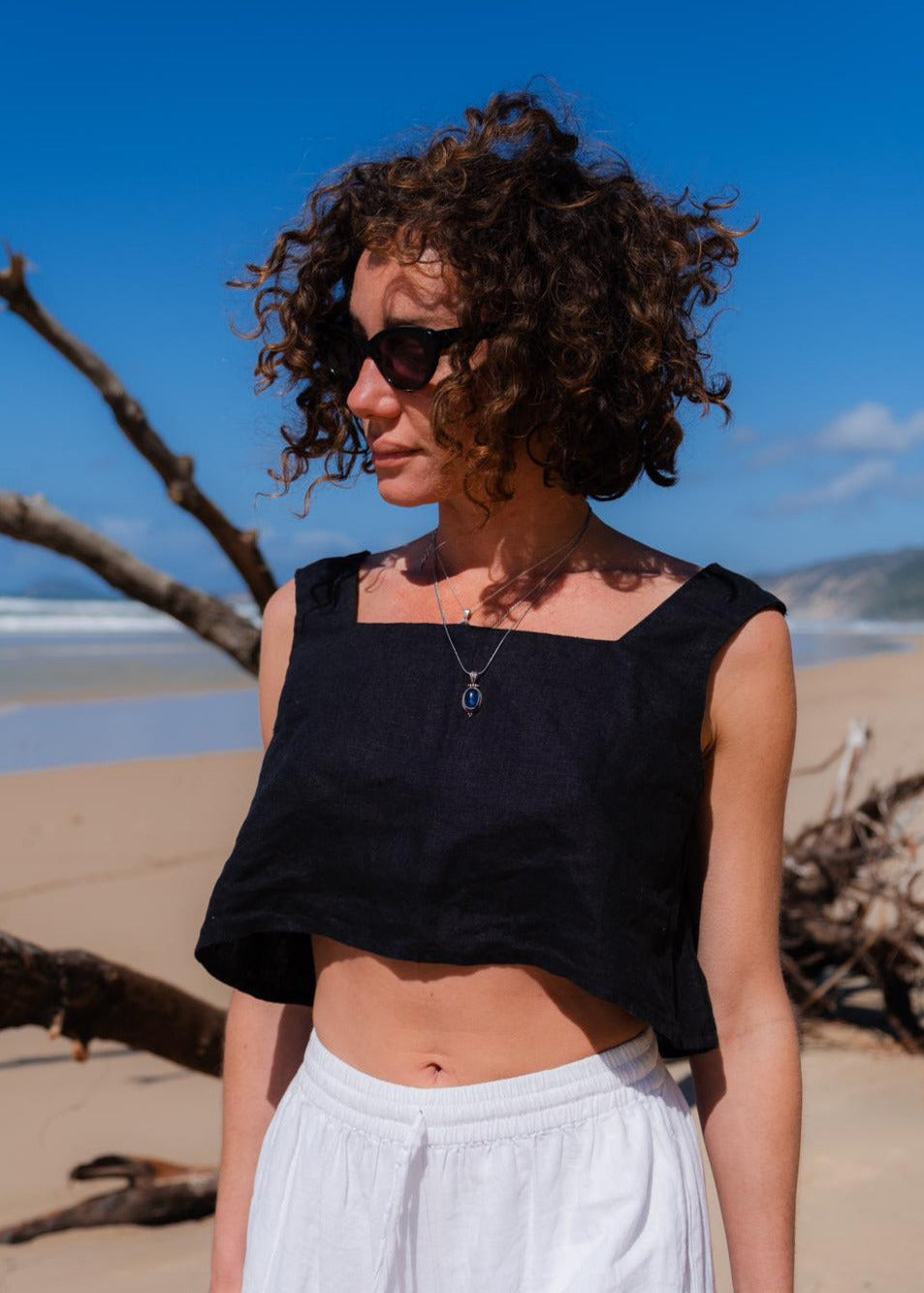 Emmie Lane standing on a beach wearing a black top and white pants with sunglasses.